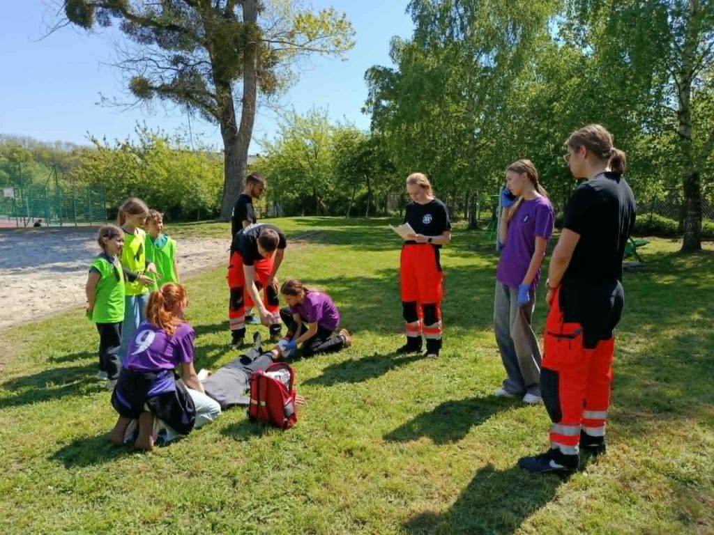 Na tle zewnętrznej siłowni trzyosobowa drużyna udzielająca pomocy chłopcu leżącemu na trawie. W koło ratownicy oceniający udzielanie pomocy oraz troje małych uczniów.
