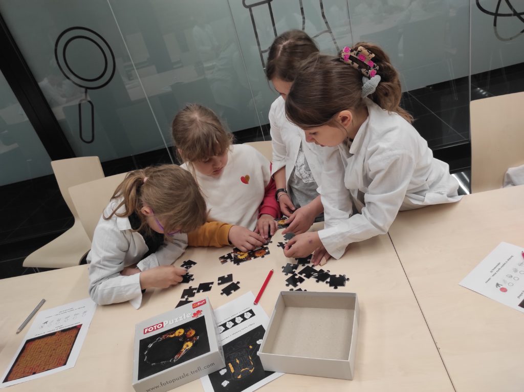 4 dzieci siedzących w fartuchach przy stole, na którym leżą puzzle i kartonik. Uczennice układają wspólnie puzzle.