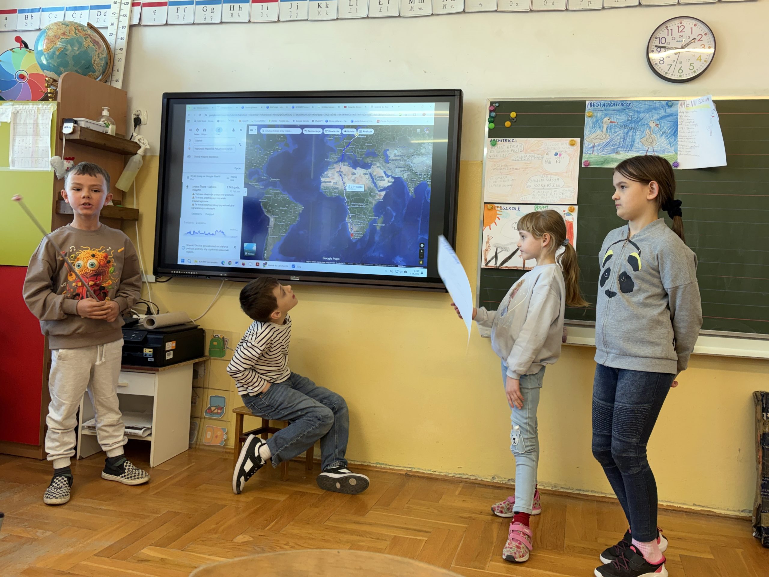 Czwórka uczniów prezentuje projekt w klasie. Na dużym ekranie wyświetlona jest mapa świata z zaznaczoną trasą migracji ptaków z Europy do Afryki, a chłopiec po lewej stronie wskazuje na nią wskaźnikiem.