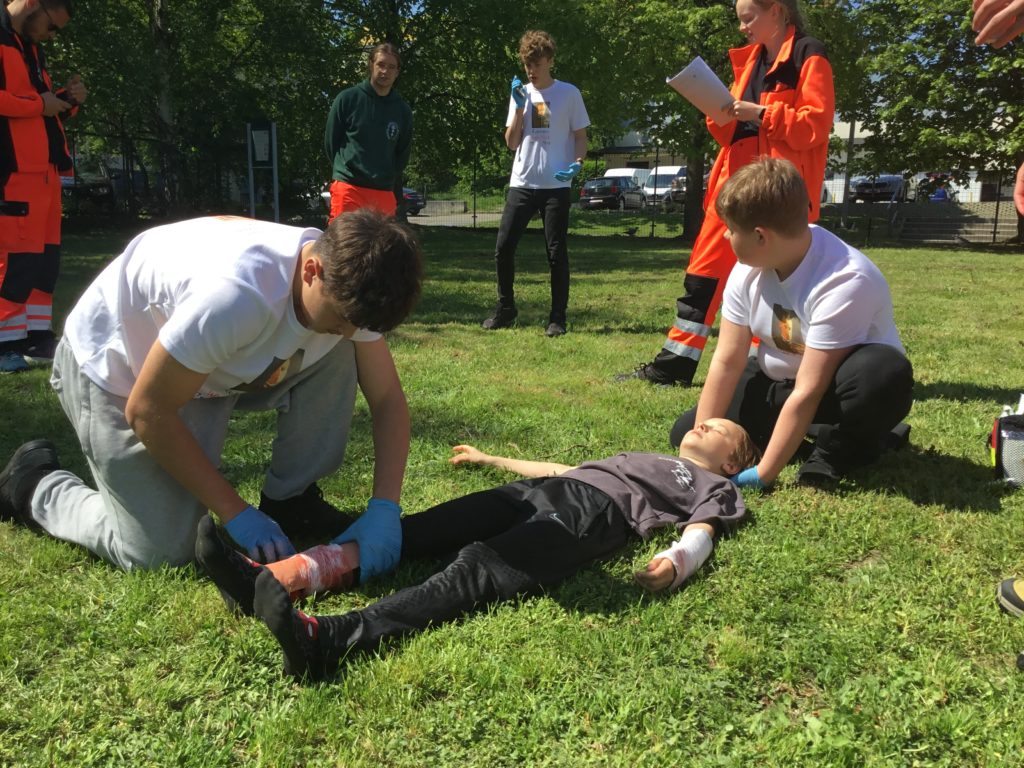 Na tle zewnętrznej siłowni trzyosobowa drużyna udzielająca pomocy chłopcu leżącemu na trawie. Jeden z nich wzywa przez telefon pomoc. W koło ratownicy oceniający udzielanie pomocy.