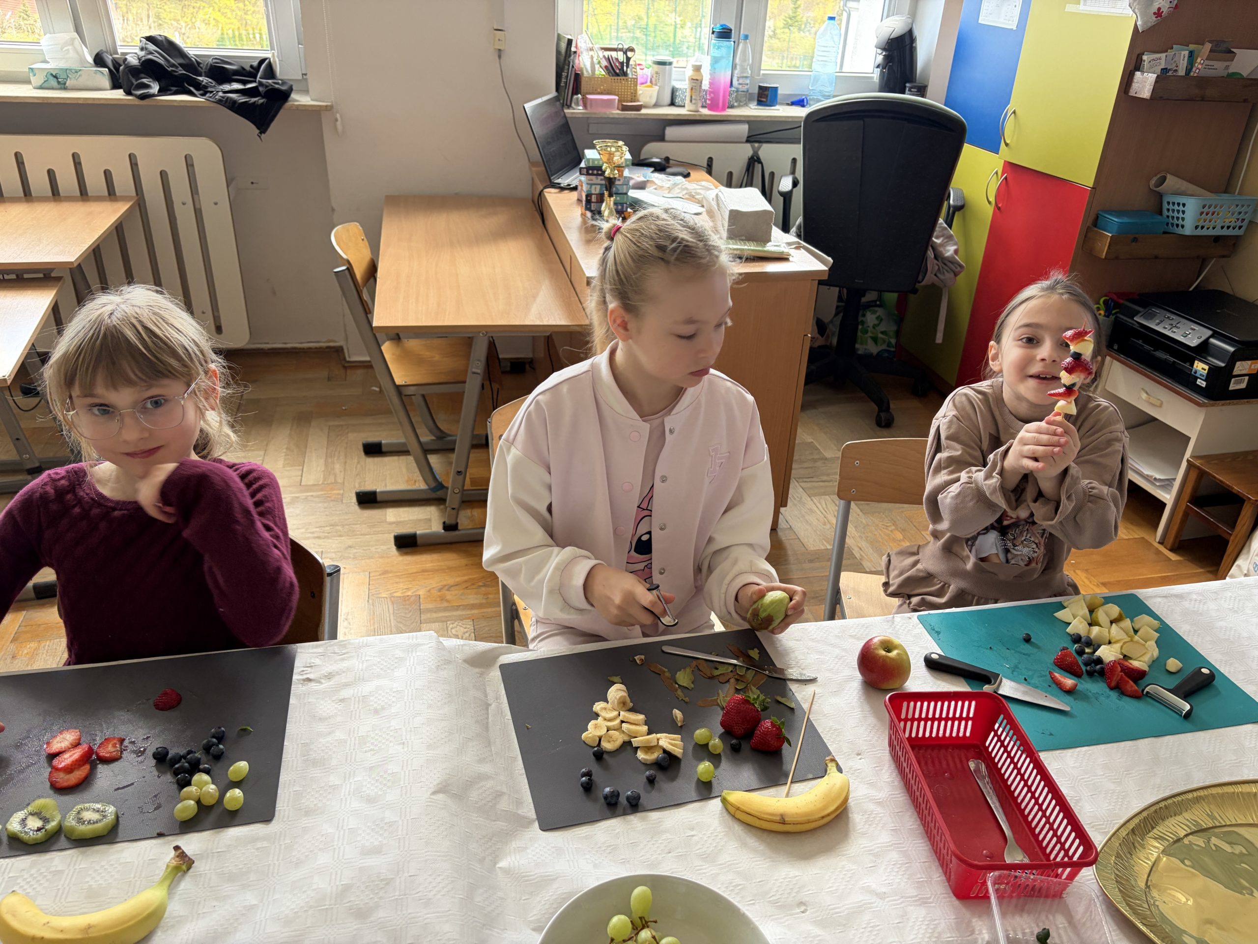Trzy dziewczynki siedzą przy stole. Dziewczynka po lewej w bordowym swetrze patrzy na przygotowane owoce (kiwi, winogrona, truskawki, borówki). Dziewczynka w środku obiera owoc obieraczką. Dziewczynka po prawej uśmiecha się i pokazuje gotowy szaszłyk owocowy. Na stole leży czerwony koszyczek z nożem i widelcem.