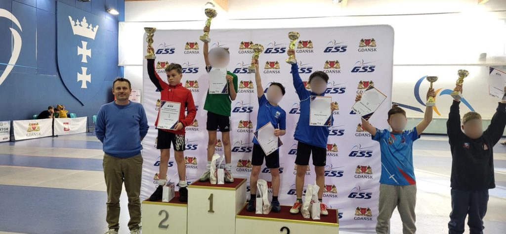 Oficjalne wręczenie nagród na hali sportowej w Gdańsku. Na tle białej ścianki sponsorskiej stoi grupa młodych zawodników w strojach sportowych oraz dorosły mężczyzna w niebieskiej bluzie. Na podium stoją trzej chłopcy: na pierwszym miejscu zawodnik w zielonej koszulce, na drugim w czerwonej bluzie, a na trzecim w niebieskiej koszulce. Obok podium stoją kolejni nagrodzeni zawodnicy. Wszyscy chłopcy dumnie wznoszą w górę złote puchary i trzymają dyplomy. Po lewej stronie widać duży, niebieski herb Gdańska na ścianie hali.