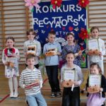 8 uczniów ustawionych w 2 rzędach, jeden siedzi, drugi stoi. Dzieci trzymają w dłoniach dyplomy i nagrody. W tle tablica informacyjna z napisem: KONKURS RECYTATORSKI „Poetycki Dzień Matki” oraz kolorowymi kwiatami, motylami i sercami. W tle elementy sali gimnastycznej.