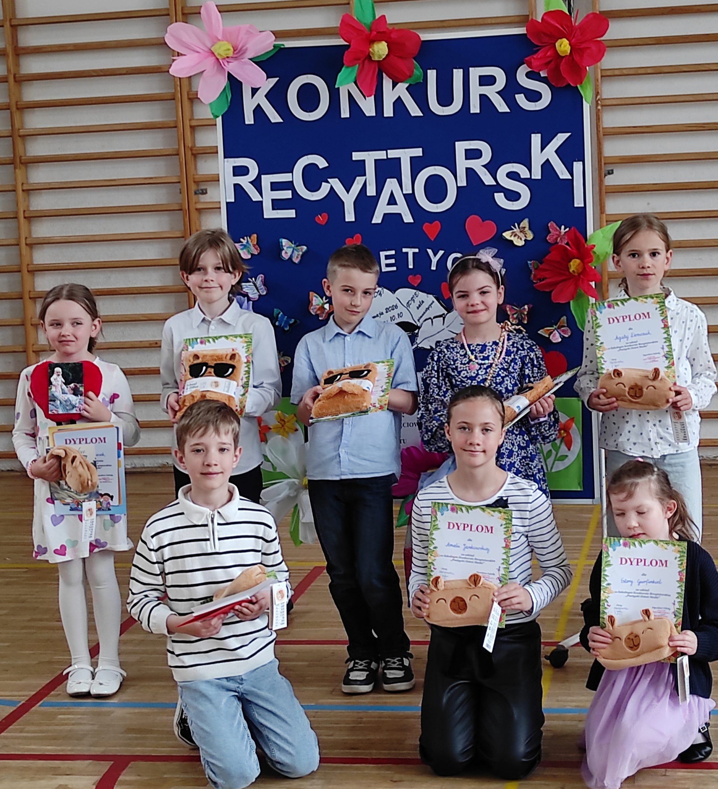 8 uczniów ustawionych w 2 rzędach, jeden siedzi, drugi stoi. Dzieci trzymają w dłoniach dyplomy i nagrody. W tle tablica informacyjna z napisem: KONKURS RECYTATORSKI „Poetycki Dzień Matki” oraz kolorowymi kwiatami, motylami i sercami. W tle elementy sali gimnastycznej.