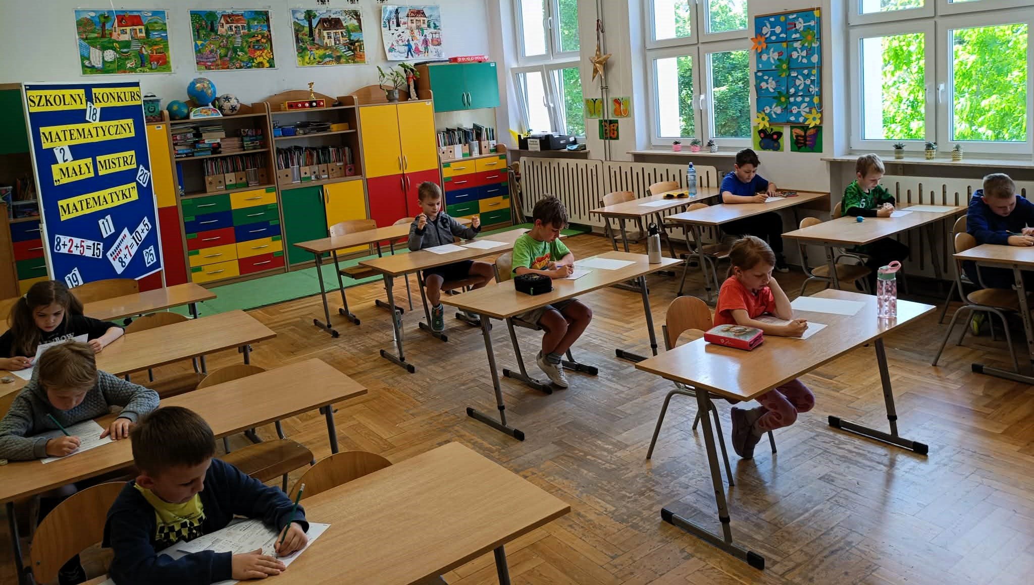 Dzieci siedzą pojedynczo w sali lekcyjnej przy ławkach i rozwiązują zadania na kartkach. Po lewej stronie znajduje się tablica z informacją o konkursie, a w tle kolorowe szafki, prace uczniów i duże okna.