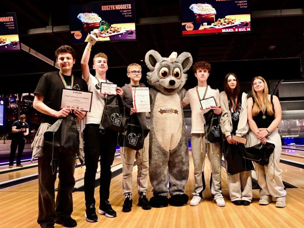 Sześcioro uczniów klas 8 stoi na torze do kręgli podczas ceremonii wręczenia nagród. Kilka osób trzyma dyplomy, medale i torby z upominkami, a jedna z nich unosi trofeum. W centrum znajduje się osoba w kostiumie szarego zwierzęcia-maskotki. W tle widoczne są tory bowlingowe oraz ekrany z reklamami.