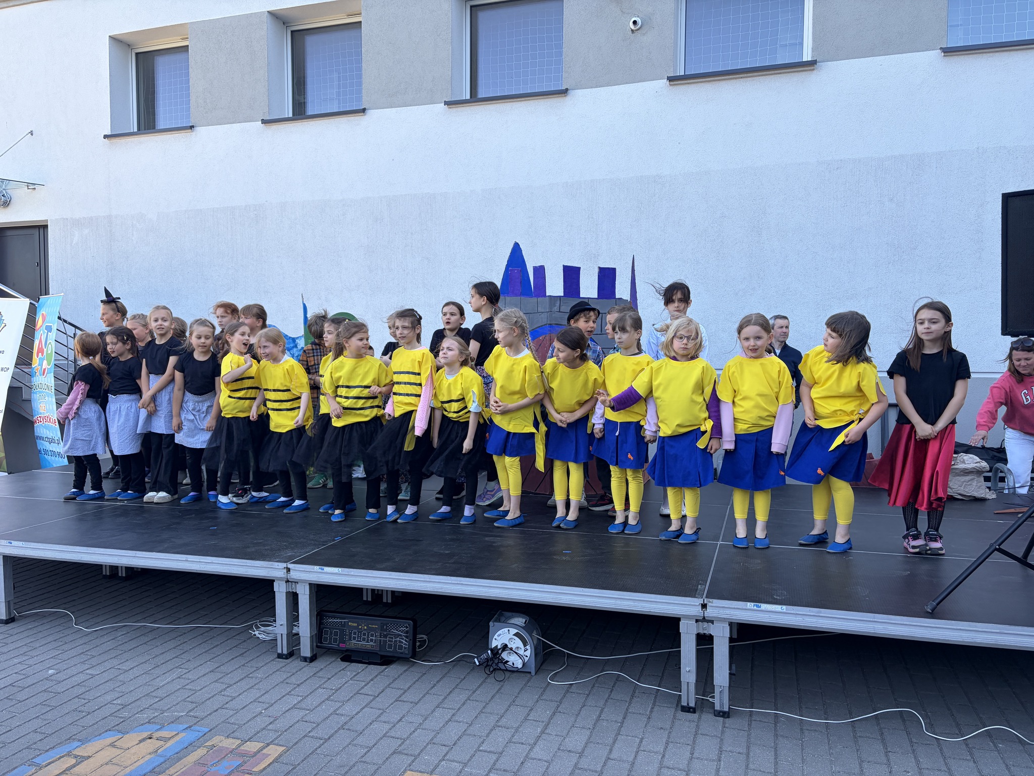 Liczna grupa dzieci w kostiumach – część w czarnych, część w żółtych koszulkach – stoi w rzędzie na scenie plenerowej, przygotowując się do występu.