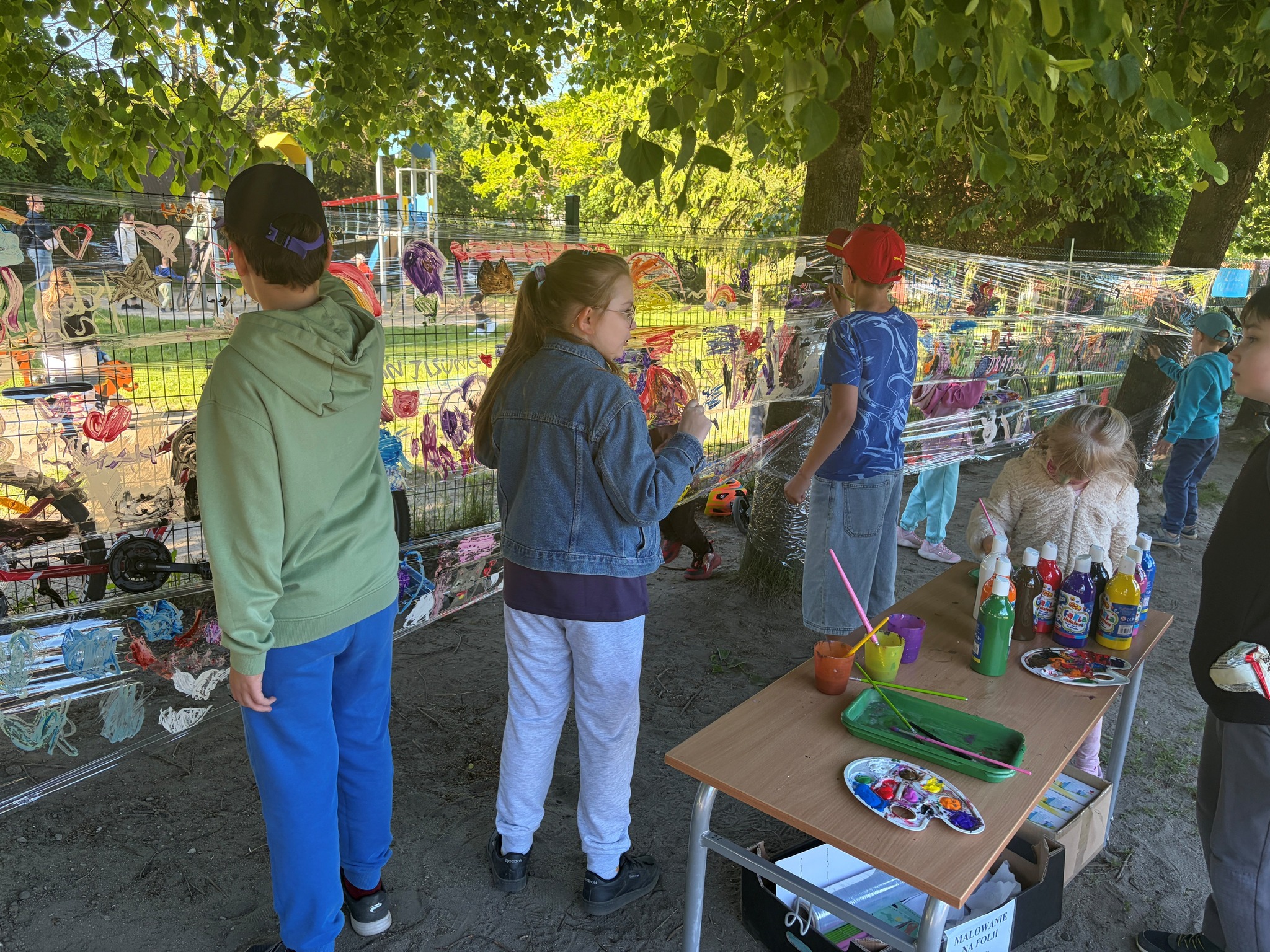 Dzieci malują farbami na przezroczystej folii rozciągniętej między drzewami. Obok stoi mały stolik z kolorowymi butelkami farb, pędzlami i plastikowymi paletami do mieszania kolorów.