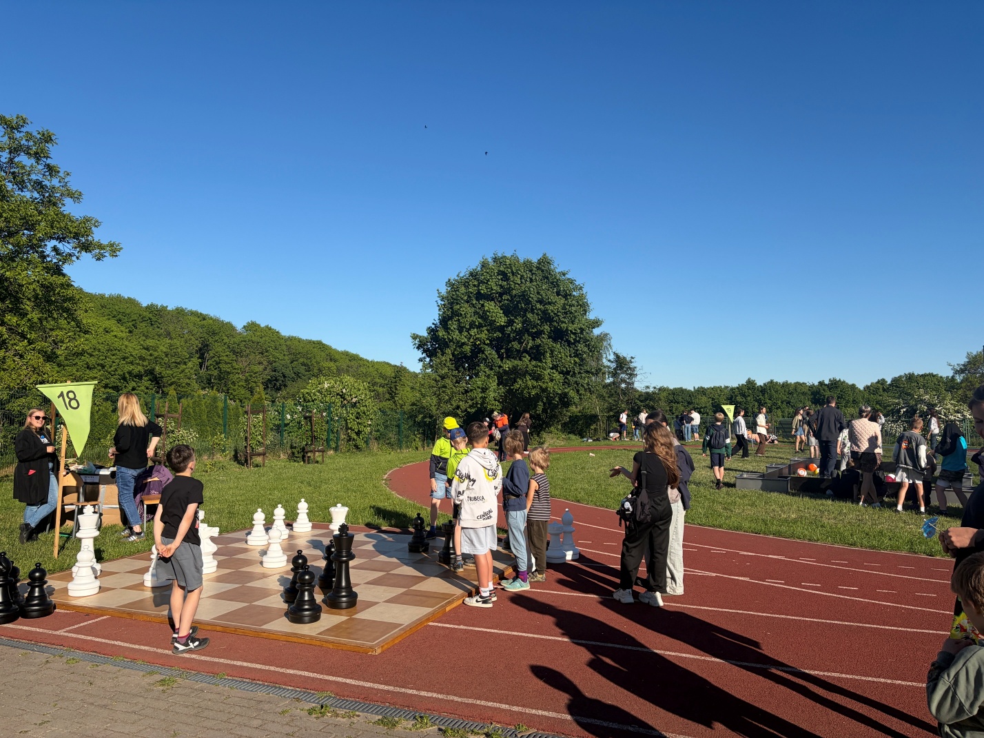 Duża plansza do gry w szachy plenerowe ułożona na tartanie obok bieżni lekkoatletycznej. Wokół dużych figur szachowych stoją dzieci i dorośli, a w tle widać zielone drzewa i uczestników pikniku.
