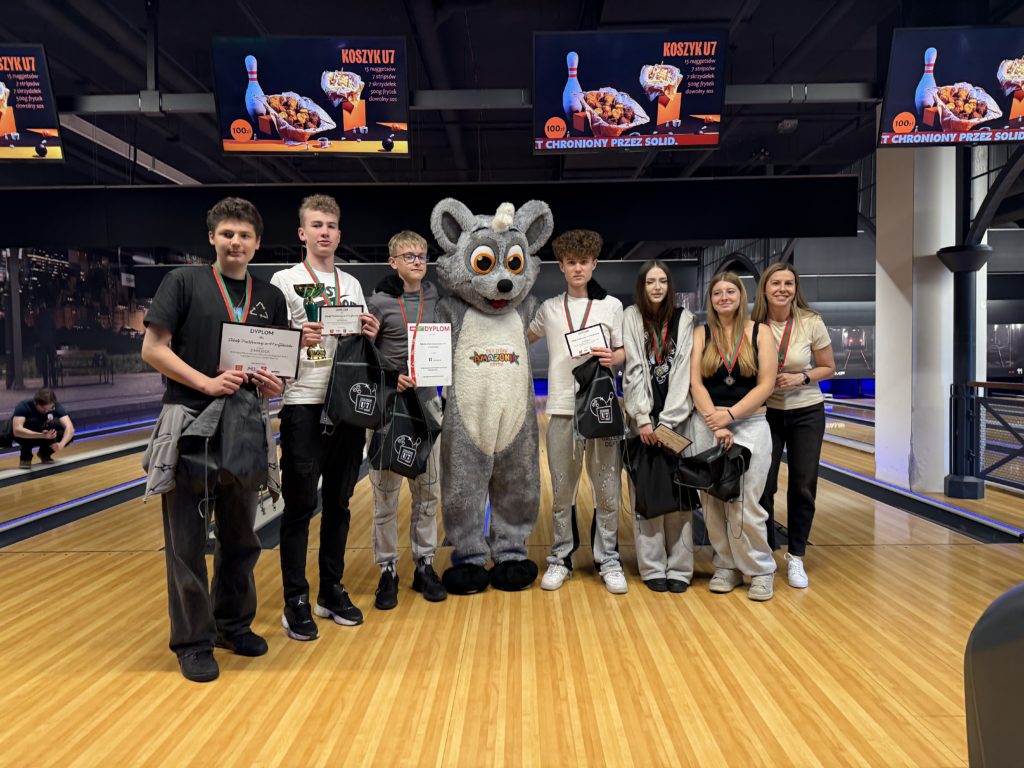 Ośmioosobowa grupa stoi na torze bowlingowym i pozuje do wspólnego zdjęcia po ceremonii wrę-czenia nagród. Kilkoro uczestników ma na szyi medale, trzyma dyplomy, puchar oraz torby z upo-minkami. W centrum znajduje się duża maskotka w kostiumie szarego zwierzęcia z dużymi oczami. Po obu stronach maskotki stoją młodzi laureaci, a po prawej stronie kobieta, nauczycielka. W tle widoczne są tory do kręgli oraz ekrany reklamowe zawieszone nad alejkami.