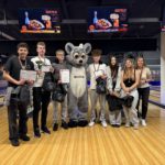 Ośmioosobowa grupa stoi na torze bowlingowym i pozuje do wspólnego zdjęcia po ceremonii wrę-czenia nagród. Kilkoro uczestników ma na szyi medale, trzyma dyplomy, puchar oraz torby z upo-minkami. W centrum znajduje się duża maskotka w kostiumie szarego zwierzęcia z dużymi oczami. Po obu stronach maskotki stoją młodzi laureaci, a po prawej stronie kobieta, nauczycielka. W tle widoczne są tory do kręgli oraz ekrany reklamowe zawieszone nad alejkami.