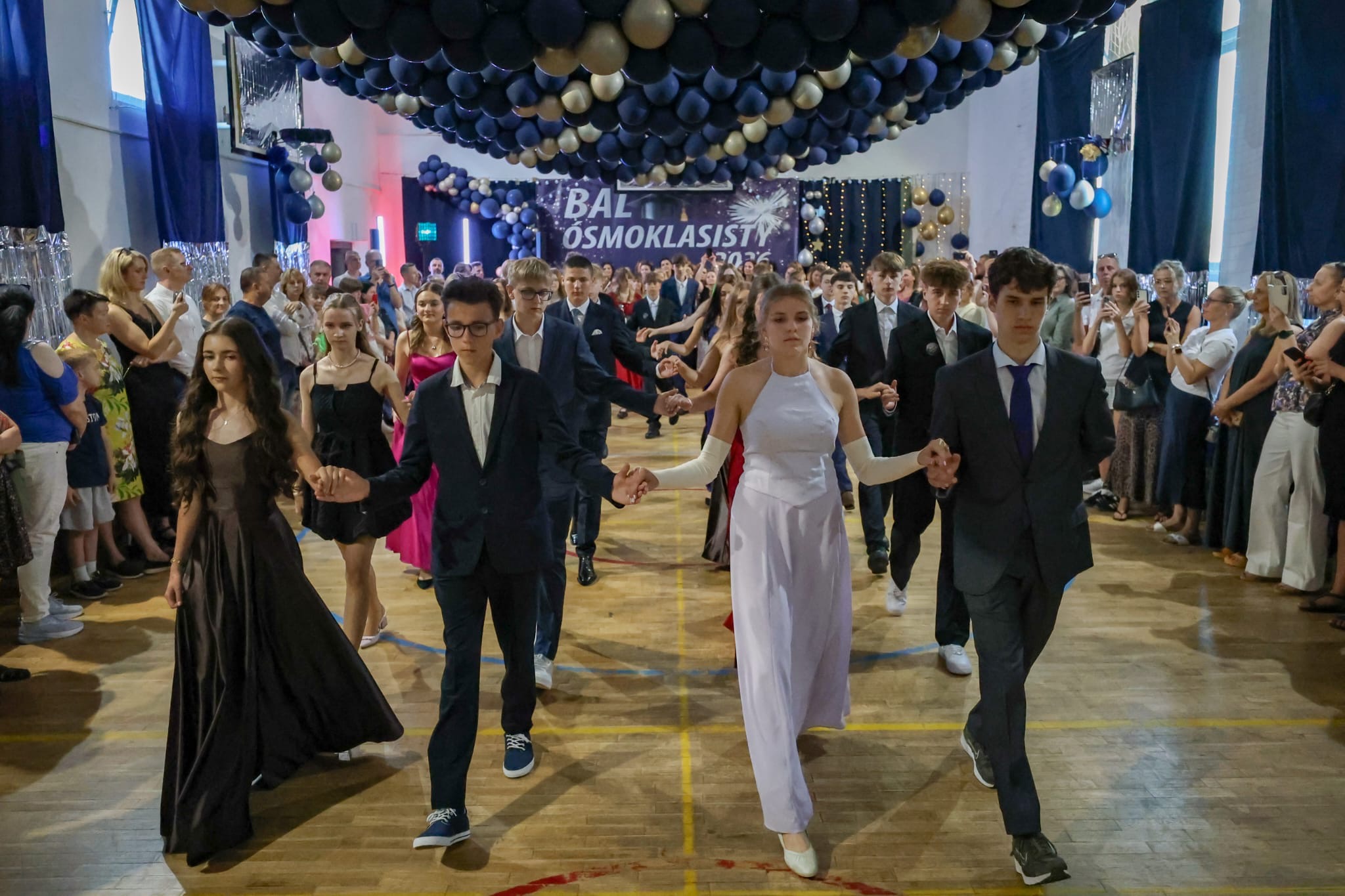 Uczniowie klas ósmych w eleganckich strojach wykonują uroczysty taniec podczas balu ósmoklasi-sty. Na pierwszym planie widać pary idące w szyku, trzymające się za ręce. Wokół parkietu stoją rodzice i goście obserwujący występ, a nad uczestnikami znajduje się efektowna dekoracja z grana-towych, czarnych i złotych balonów. W tle widoczny jest baner z napisem „Bal Ósmoklasisty 2026”, podkreślający odświętny charakter wydarzenia.