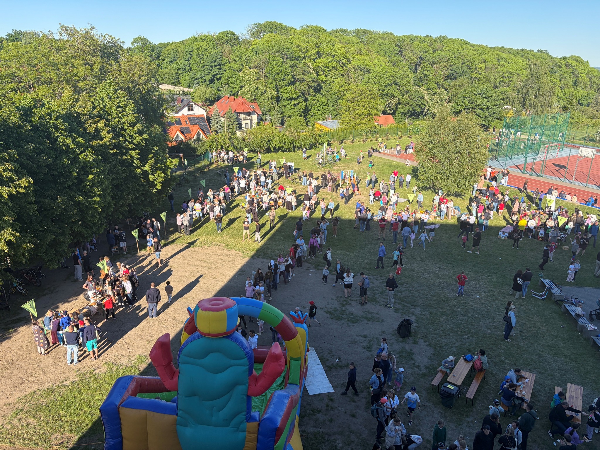 Piknik plenerowy na trawiastym terenie z lotu ptaka. Na obrazie widać tłum ludzi, dmuchany zamek, stoiska i boisko sportowe otoczone drzewami i domami w tle.