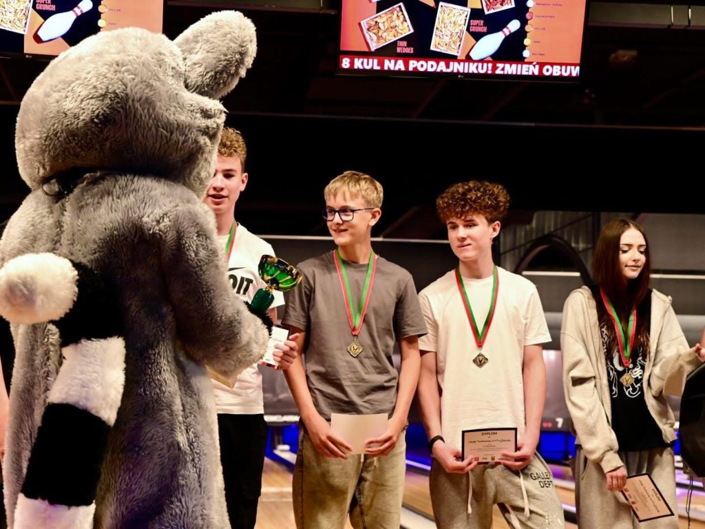 Na torze bowlingowym czworo uczestników ceremonii wręczenia nagród stoi obok dużej maskotki w kostiumie szarego zwierzęcia, widocznej od tyłu. Uczestnicy mają na szyjach medale i trzymają dyplomy lub nagrody; jedna osoba częściowo zasłonięta przez maskotkę trzyma puchar. W tle znaj-dują się tory do kręgli oraz ekrany reklamowe zawieszone nad alejkami.