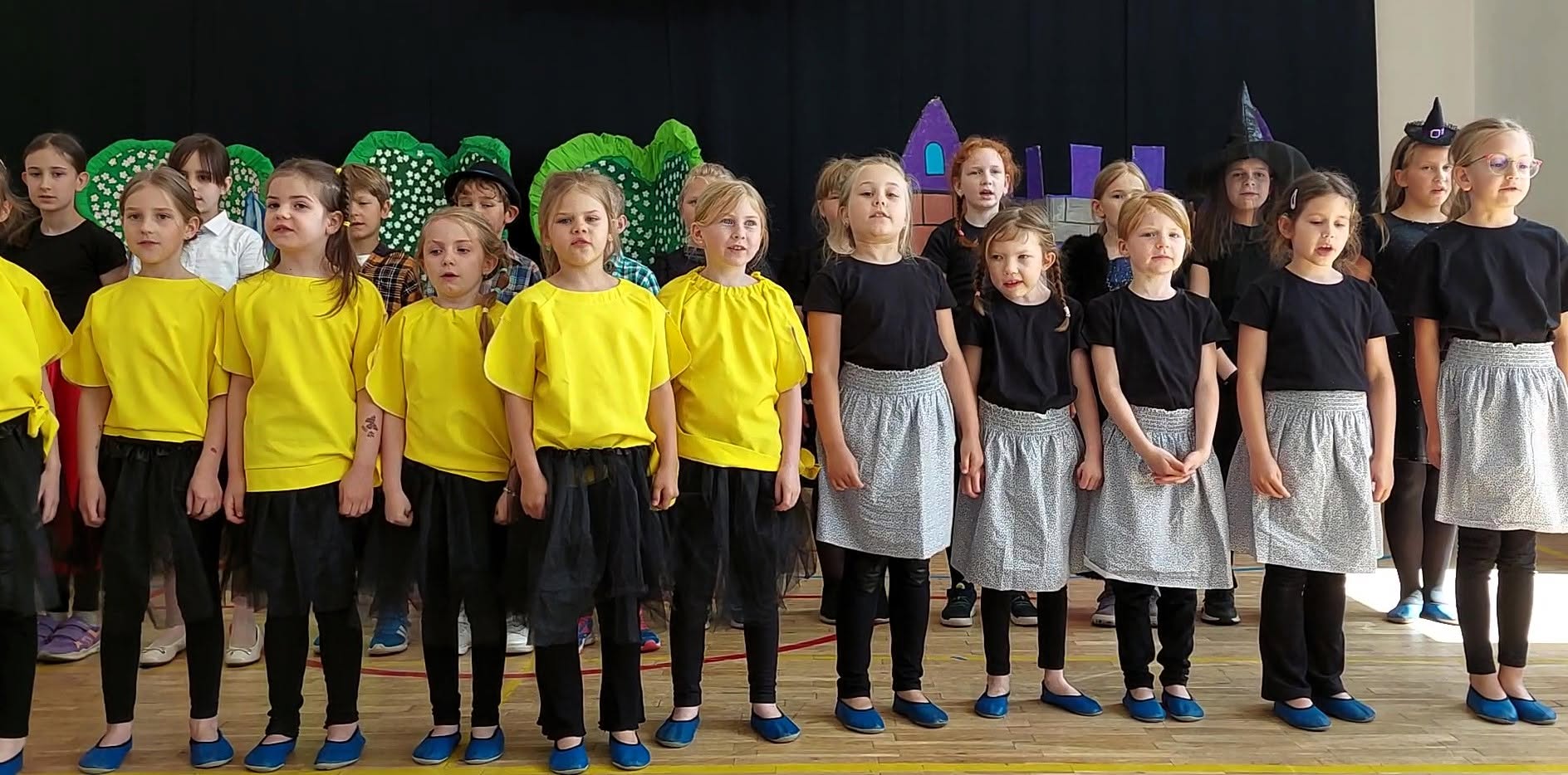 Grupa dzieci stoi na szkolnej scenie podczas przedstawienia, dzieci w kostiumach w kolorach żółtym i czarnym. W tle widać dekoracje teatralne .