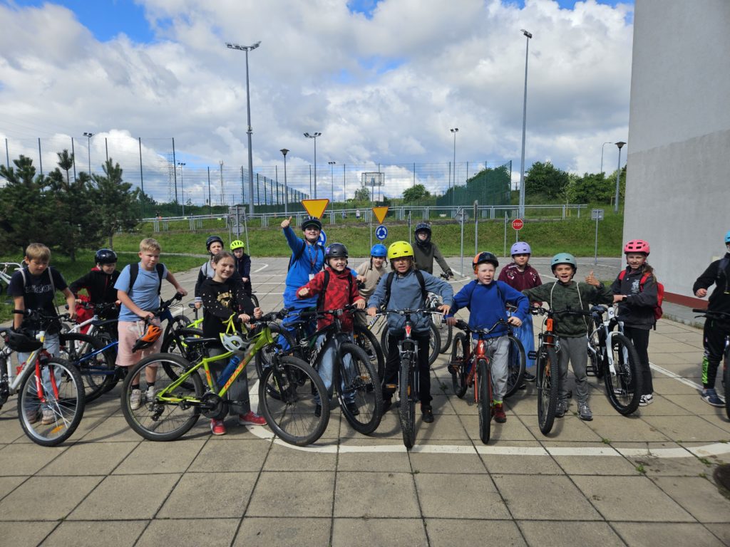 Grupka pierwszych, szczęśliwych rowerzystów ze zdanego egzaminu. Stoją na placu trzymając swoje rowery. Na głowach mają kolorowe kaski
