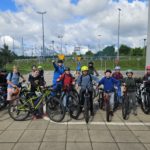 Grupka pierwszych, szczęśliwych rowerzystów ze zdanego egzaminu. Stoją na placu trzymając swoje rowery. Na głowach mają kolorowe kaski
