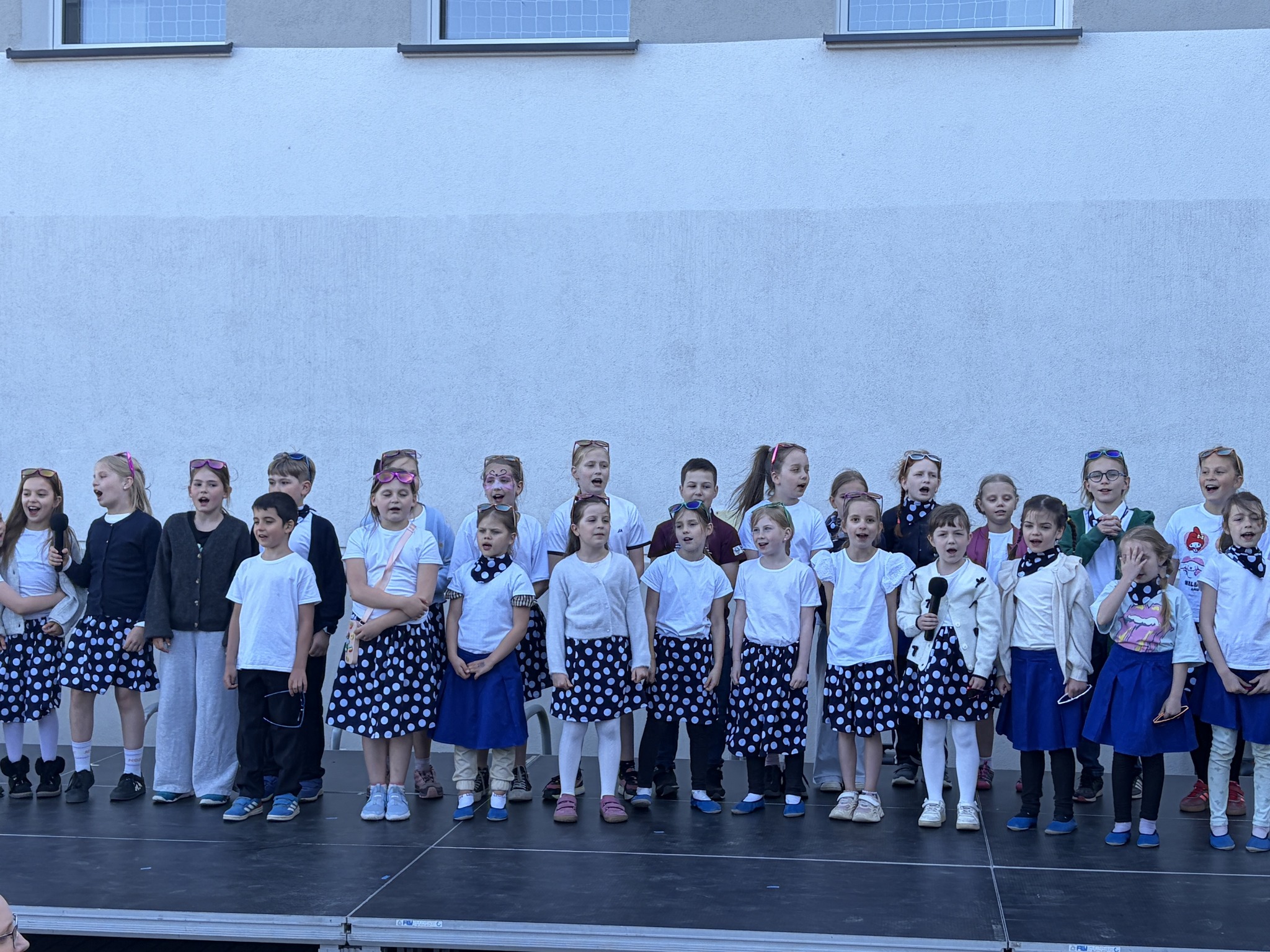 Duża grupa dzieci stojąca w rzędzie na podwyższeniu na tle jasnej ściany budynku. Dzieci są ubrane w białe koszulki oraz ciemne spódnice i spodnie, wspólnie śpiewają do mikrofonów podczas szkolnego występu.