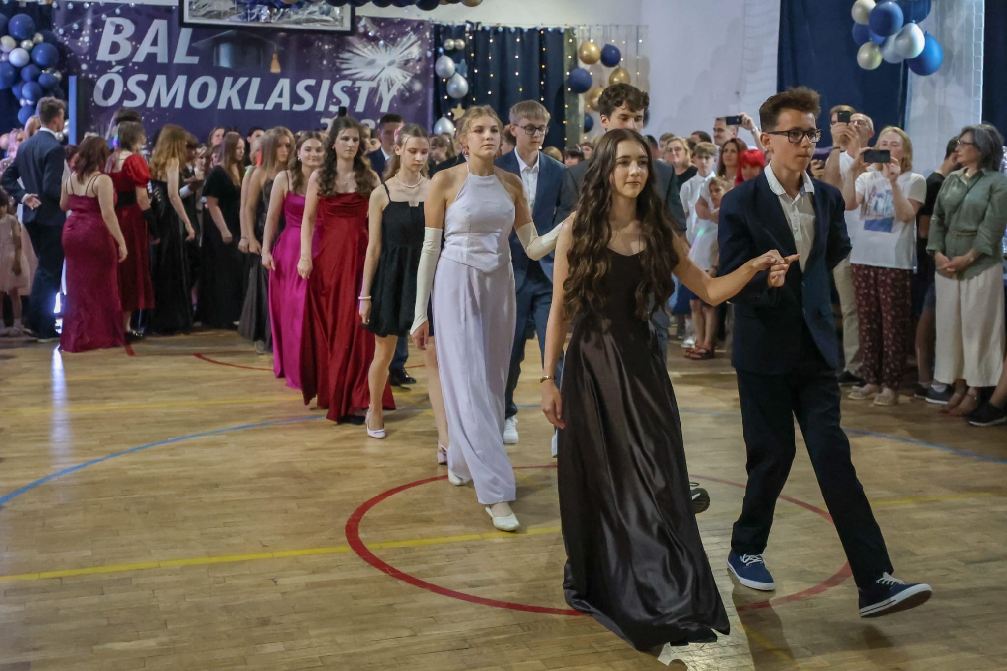 Uczniowie klasy ósmej w strojach galowych wchodzą parami na parkiet podczas balu ósmoklasisty, otoczeni przez zgromadzonych gości i dekoracje balowe.