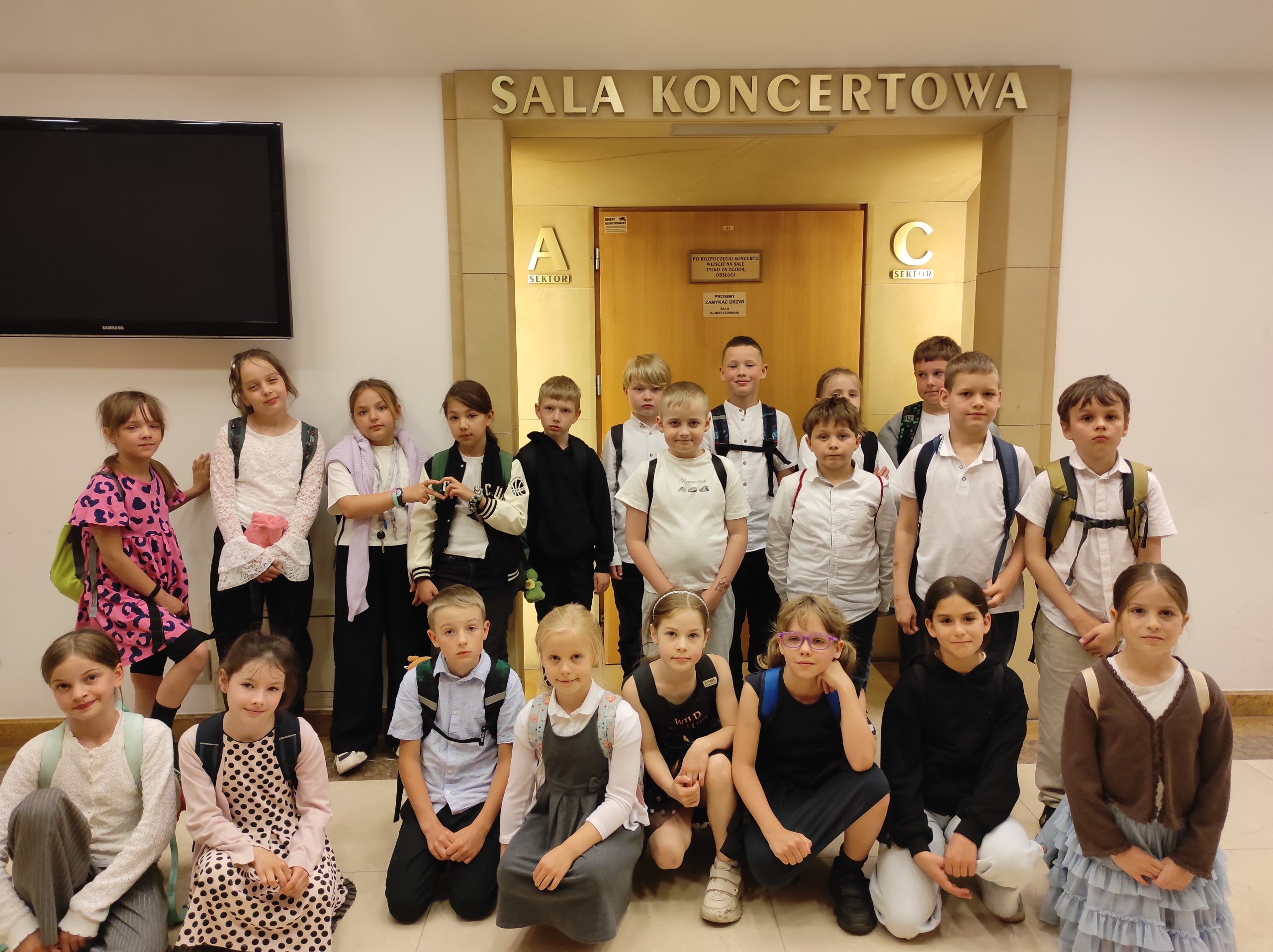 21 dzieci ustawionych w dwa rzędy, jeden stoi, drugi kuca. W tle fragment ściany oraz napis SALA KONCERTOWA.
