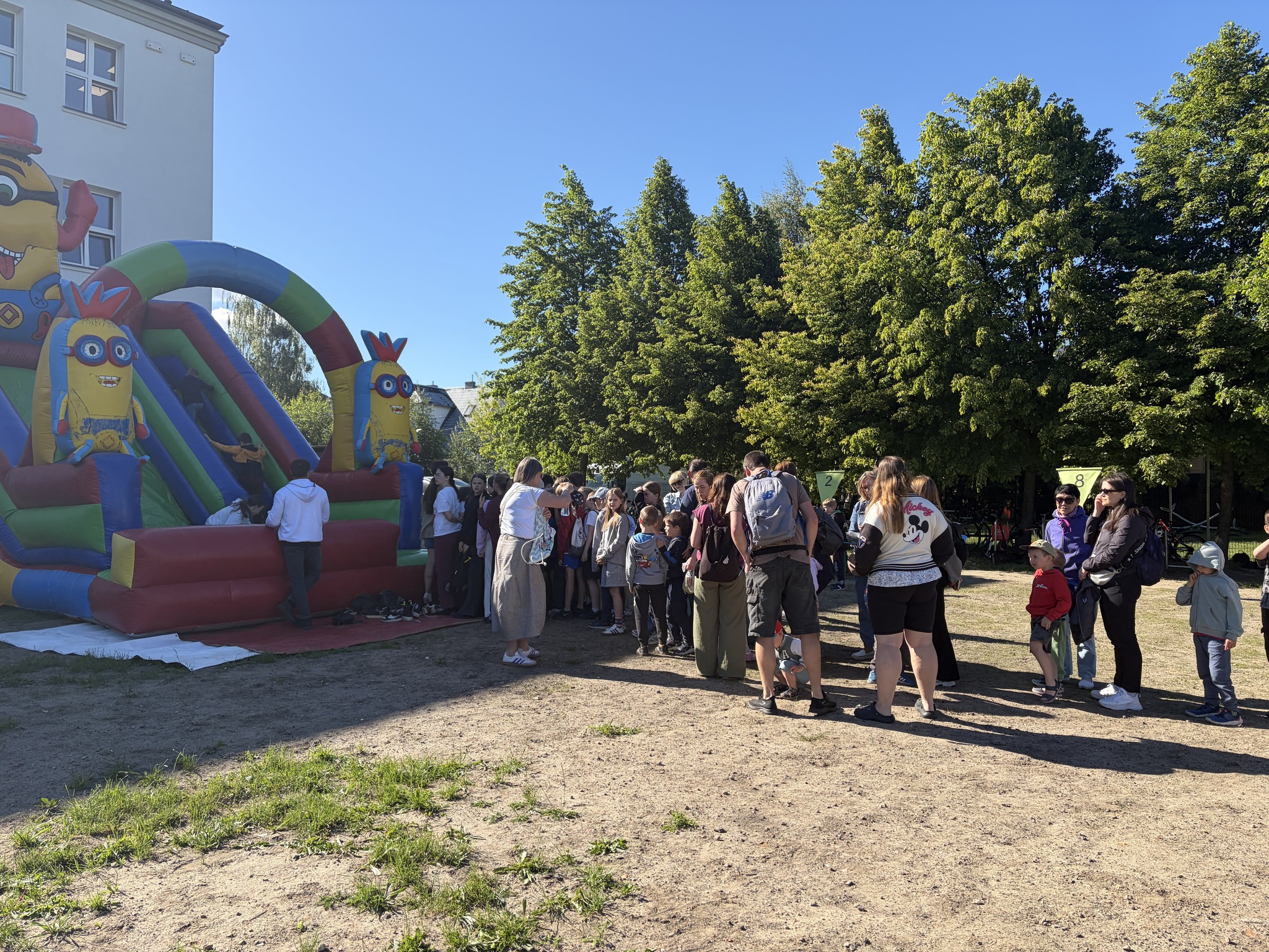 Kolejka dzieci i rodziców czekających na trawiastym placu obok budynku szkoły. Po lewej stronie stoi duża, kolorowa dmuchana zjeżdżalnia z motywem Minionków. W tle rosną wysokie, zielone drzewa.