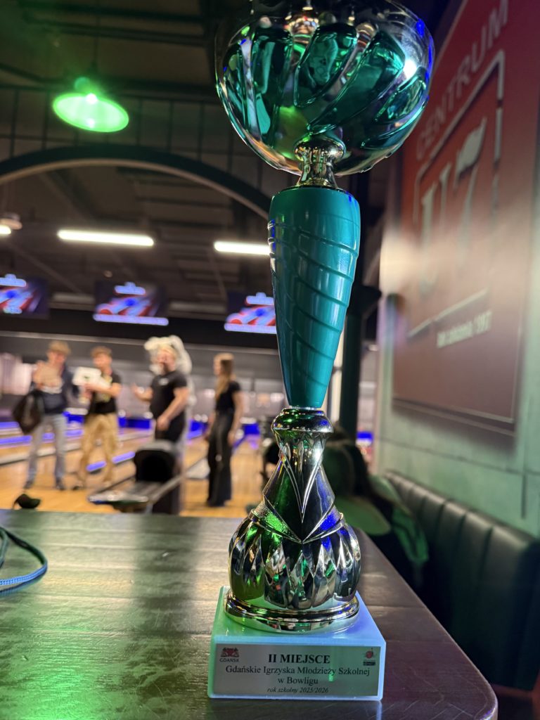 Srebrno-turkusowy puchar stoi na stole w kręgielni. Na tabliczce widnieje napis: „II MIEJSCE - Gdańskie Igrzyska Młodzieży Szkolnej w Bowlingu, rok szkolny 2025/2026”. W tle widać rozmyte tory bowlingowe oraz kilka osób.