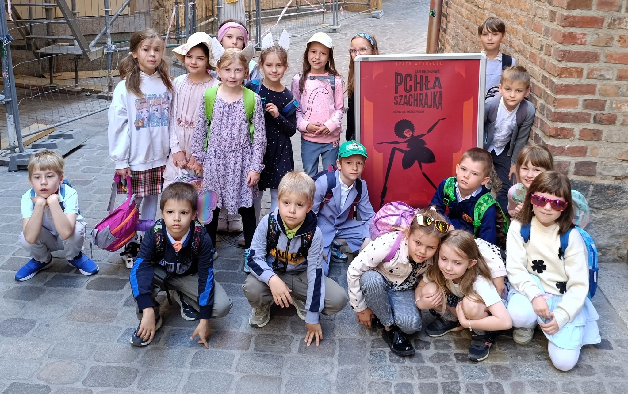 Grupa kilkunastu dzieci w wieku szkolnym pozuje na brukowanej uliczce między ceglanymi murami. Większość ma plecaki, a niektóre noszą czapki, opaski lub okulary przeciwsłoneczne. W centrum stoi czerwony plakat teatralny z napisem „Pchła Szachrajka”. Dzieci stoją i kucają wokół plakatu, patrząc w stronę aparatu. W tle widoczne są rusztowania i ogrodzenie remontowanego budynku.