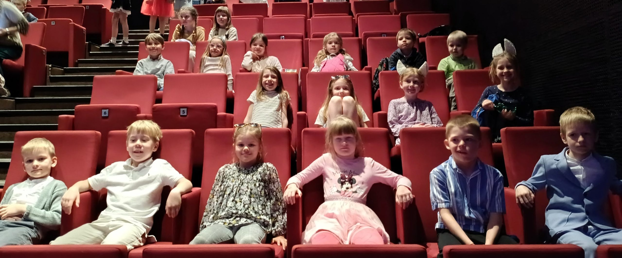 Grupa dzieci siedzi w rzędach czerwonych foteli na widowni sali teatralnej lub kinowej. Dzieci są rozmieszczone na kilku poziomach widowni i patrzą w stronę aparatu. Niektóre uśmiechają się, a część ma przy sobie plecaki lub ozdoby we włosach. W tle widoczne są kolejne rzędy czerwonych siedzeń oraz schody prowadzące między sektorami widowni. Atmosfera sugeruje oczekiwanie na rozpoczęcie przedstawienia lub seansu.