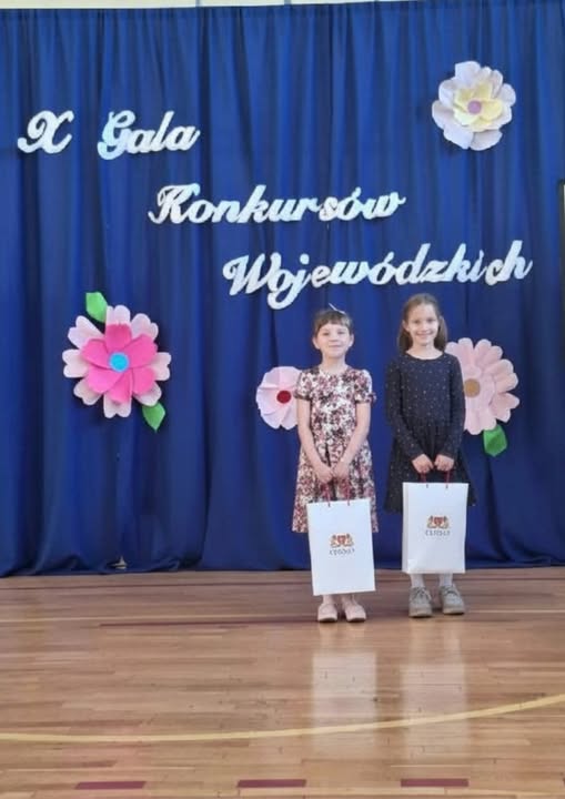 Dwie uczennice stoją na szkolnej scenie przed niebieską kurtyną z napisem ‘X Gala Konkursów Wojewódzkich’. Trzymają torby z nagrodami i uśmiechają się do zdjęcia. W tle dekoracje z papierowych kwiatów.