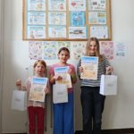 Na szkolnym korytarzu na tle tablicy z ogłoszeniami na temat zdrowia stoją trzy uczennice z klas młodszych. W rękach trzymają dyplomy i torebki z nagrodami.