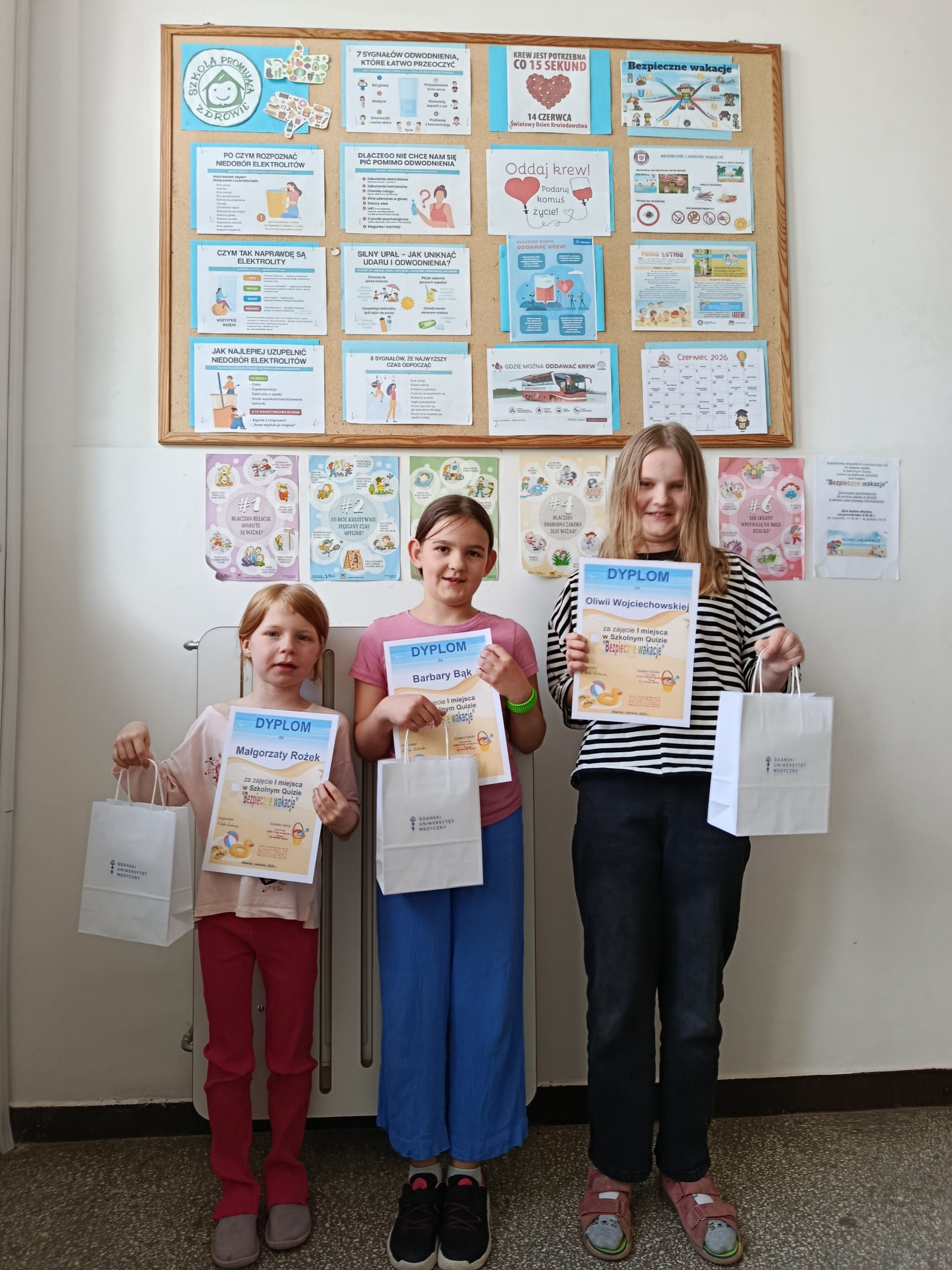 Na szkolnym korytarzu na tle tablicy z ogłoszeniami na temat zdrowia stoją trzy uczennice z klas młodszych. W rękach trzymają dyplomy i torebki z nagrodami.