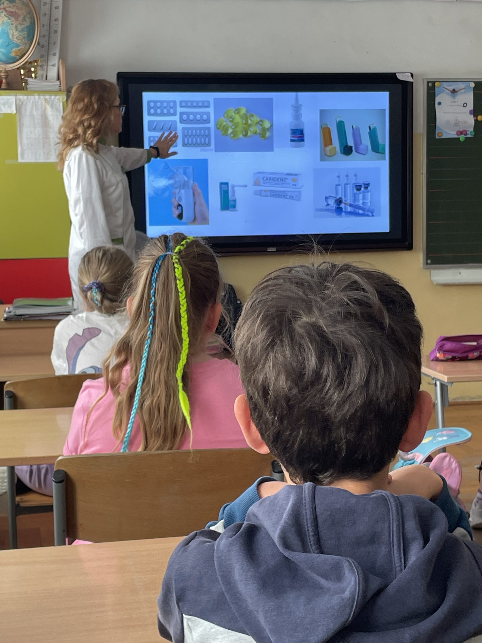 Pani farmaceutka pokazuje uczniom siedzącym w ławkach na tablicy multimedialnej rodzaje leków.