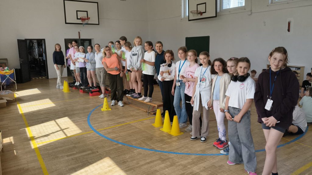 Grupa uczniów klas 4 i trzech dorosłych stoi w rzędzie w szkolnej sali gimnastycznej. Uczestnicy ustawieni są na ławkach i niskich podestach między żółtymi pachołkami. Niektóre dzieci trzymają dyplomy i bidony, a część ma na szyi identyfikatory lub medale. W tle widać kosze do koszykówki, wysokie okna oraz inne osoby siedzące przy ścianie.