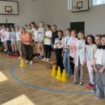 Grupa uczniów klas 4 i trzech dorosłych stoi w rzędzie w szkolnej sali gimnastycznej. Uczestnicy ustawieni są na ławkach i niskich podestach między żółtymi pachołkami. Niektóre dzieci trzymają dyplomy i bidony, a część ma na szyi identyfikatory lub medale. W tle widać kosze do koszykówki, wysokie okna oraz inne osoby siedzące przy ścianie.