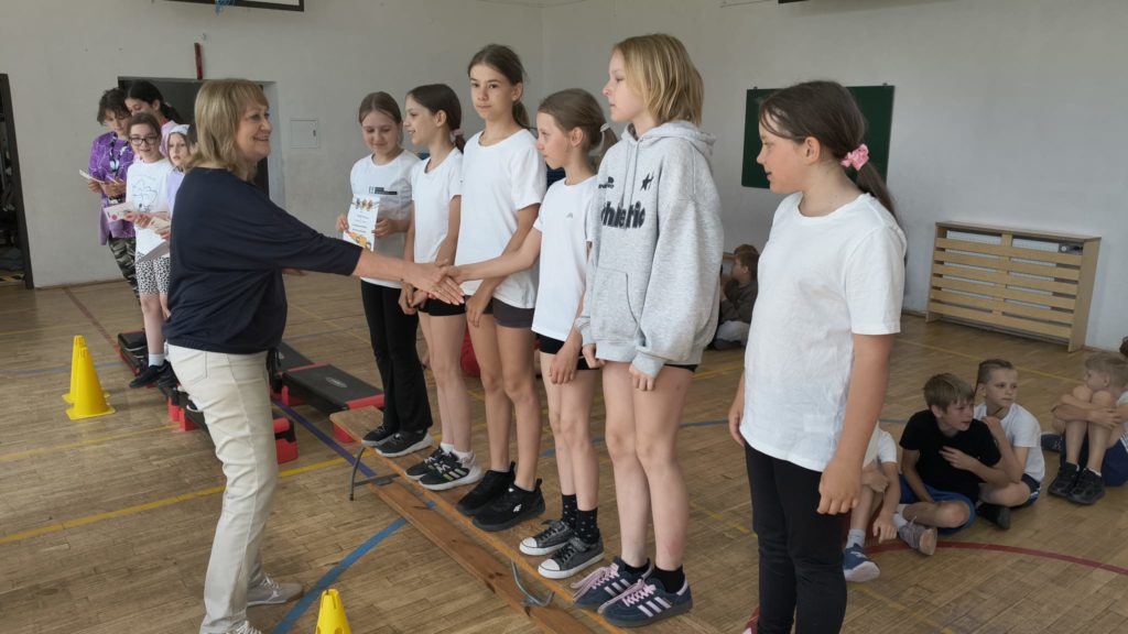 W szkolnej sali gimnastycznej pani dyrektor szkoły wręcza gratulacje i ściska dłoń jednej z uczennic stojących w rzędzie na ławce gimnastycznej. Obok znajduje się kilka dziewcząt ubranych w stroje sportowe, a jedna z nich trzyma dyplom. W tle widać innych uczniów obserwujących wydarzenie, żółte pachołki wyznaczające tor oraz drewnianą podłogę sali.
