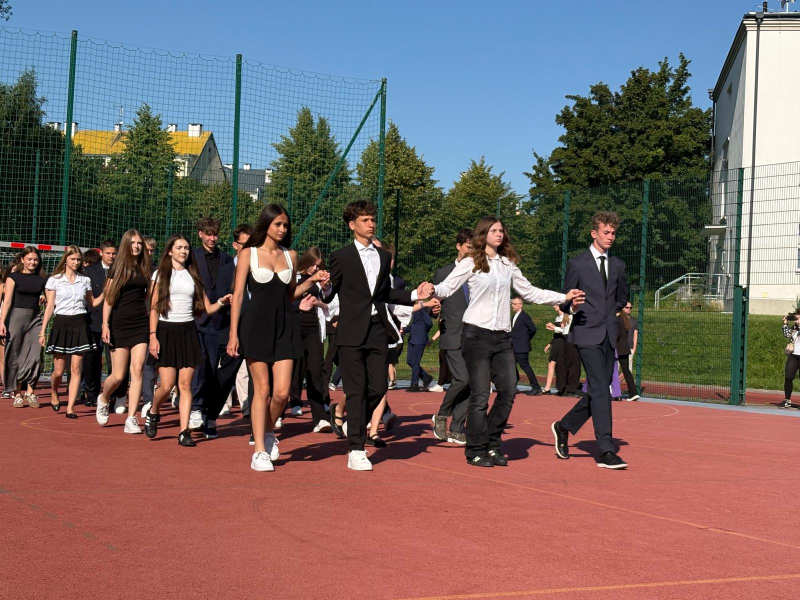 Grupa elegancko ubranych uczniów klas 8 idzie czwórkami po szkolnym boisku podczas uroczystości zakończenia roku szkolnego. W tle widać ogrodzenie boiska, drzewa oraz budynki szkolne.