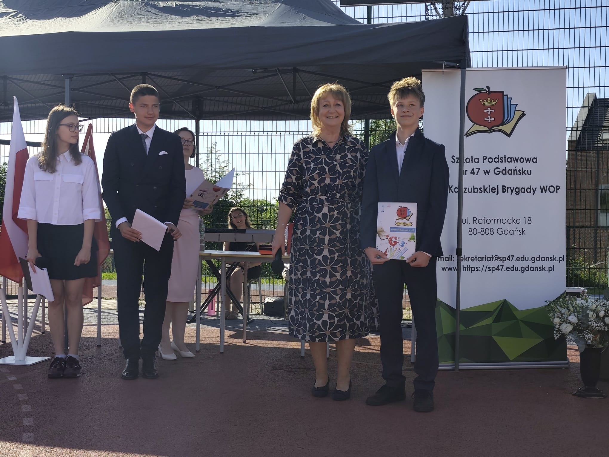 Na szkolnym boisku trwa uroczystość zakończenia roku szkolnego. Na pierwszym planie pani dyrektor stoi obok ucznia w granatowym garniturze, który trzyma dyplom. Po lewej stronie stoją pozostali uczniowie w odświętnych strojach z dokumentami w dłoniach. W tle widoczne jest zadaszenie organizatora imprezy, baner Szkoły Podstawowej nr 47 w Gdańsku oraz polskie flagi.