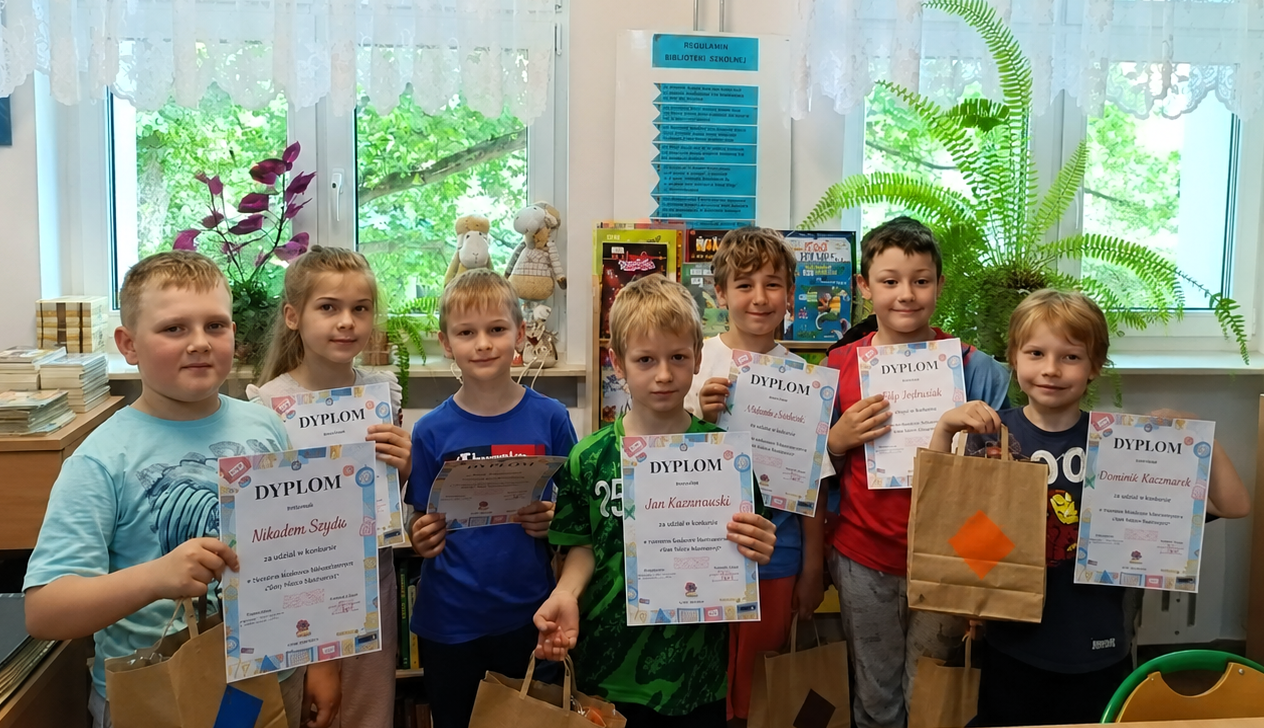 Grupa dzieci stoi w szkolnej bibliotece, trzymając dyplomy i papierowe torby z upominkami. W tle znajdują się książki, rośliny doniczkowe i okna wpuszczające naturalne światło.