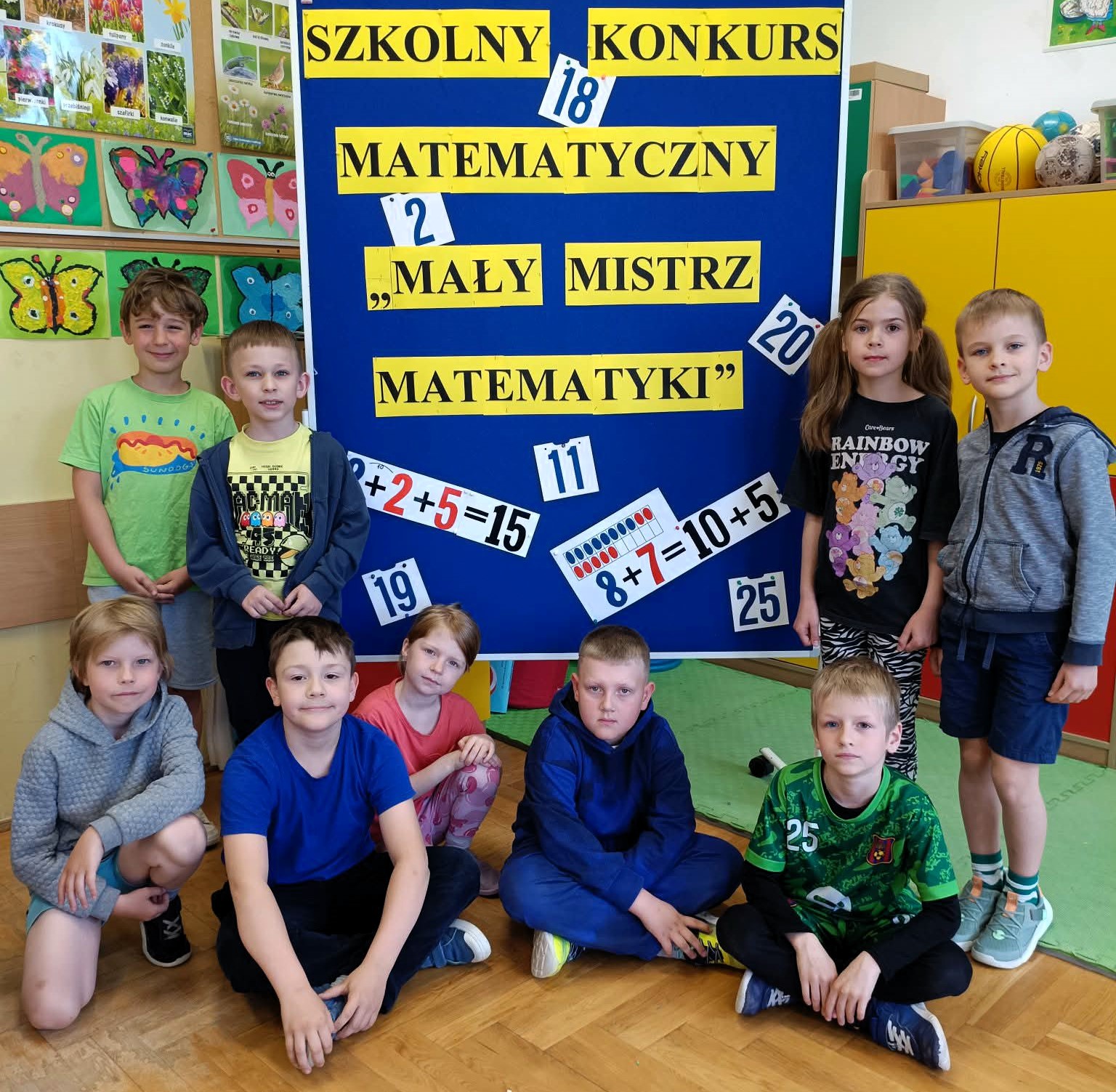 Grupa dzieci w klasie pozuje do zdjęcia przed dużą niebieską tablicą informującą o szkolnym konkursie matematycznym „Mały Mistrz Matematyki”. Na tablicy znajdują się liczby i proste działania matematyczne. W tle widoczne są kolorowe prace plastyczne przedstawiające motyle.