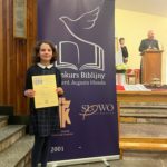 Dziewczynka stoi w kościele obok plakatu konkursu biblijnego i trzyma w rękach dyplom. W tle widoczna jest scena z duchownym przemawiającym przy mównicy