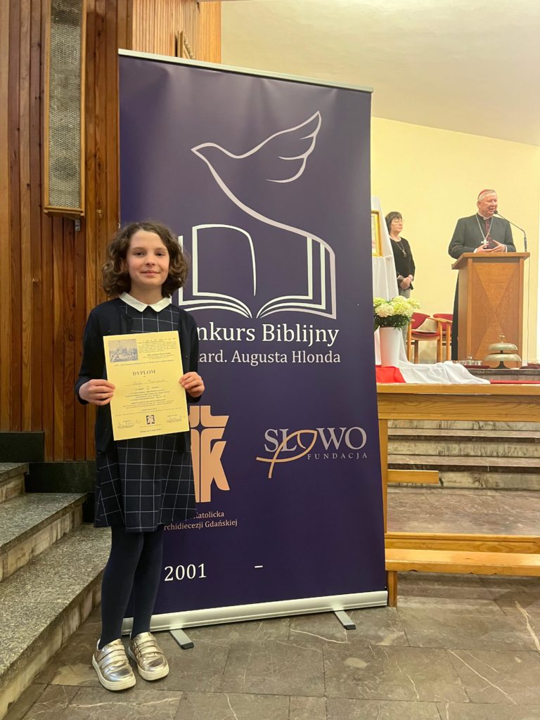 Dziewczynka stoi w kościele obok plakatu konkursu biblijnego i trzyma w rękach dyplom. W tle widoczna jest scena z duchownym przemawiającym przy mównicy