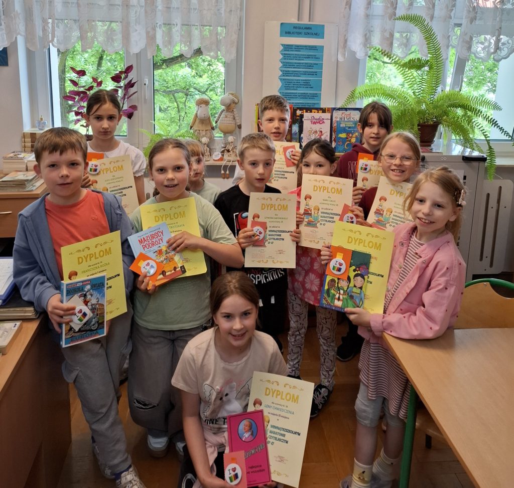 Uczniowie, dziewczynki i chłopcy stoją uśmiechnięci w pomieszczeniu biblioteki na tle okien. Każdy uczeń trzyma w ręku pamiątkowy dyplom i upominek, a osoby wyróżnione książkę.