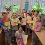 Uczniowie, dziewczynki i chłopcy stoją uśmiechnięci w pomieszczeniu biblioteki na tle okien. Każdy uczeń trzyma w ręku pamiątkowy dyplom i upominek, a osoby wyróżnione książkę.