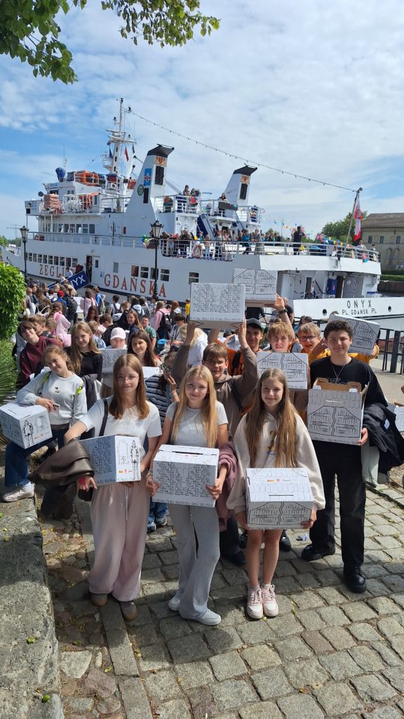 Uczniowie pozują do wspólnego zdjęcia na nabrzeżu przed statkiem udekorowanym flagami i balonami. W rękach trzymają pudełka z prezentami otrzymanymi podczas wydarzenia. W tle jest statek oraz liczni uczestnicy rejsu