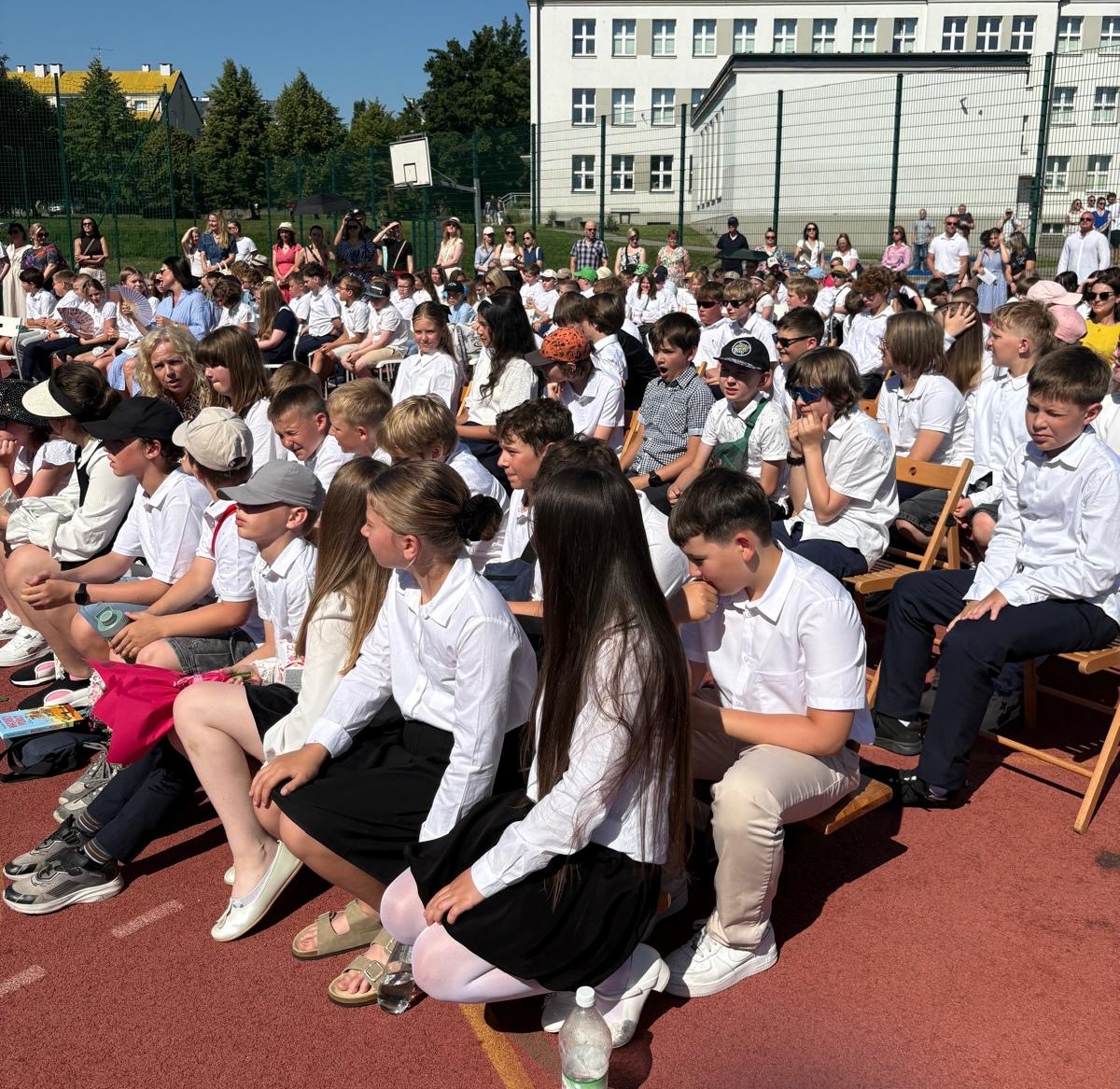  Uczniowie i nauczyciele siedzą na krzesłach na szkolnym boisku, uczestnicząc w uroczystości na świeżym powietrzu.