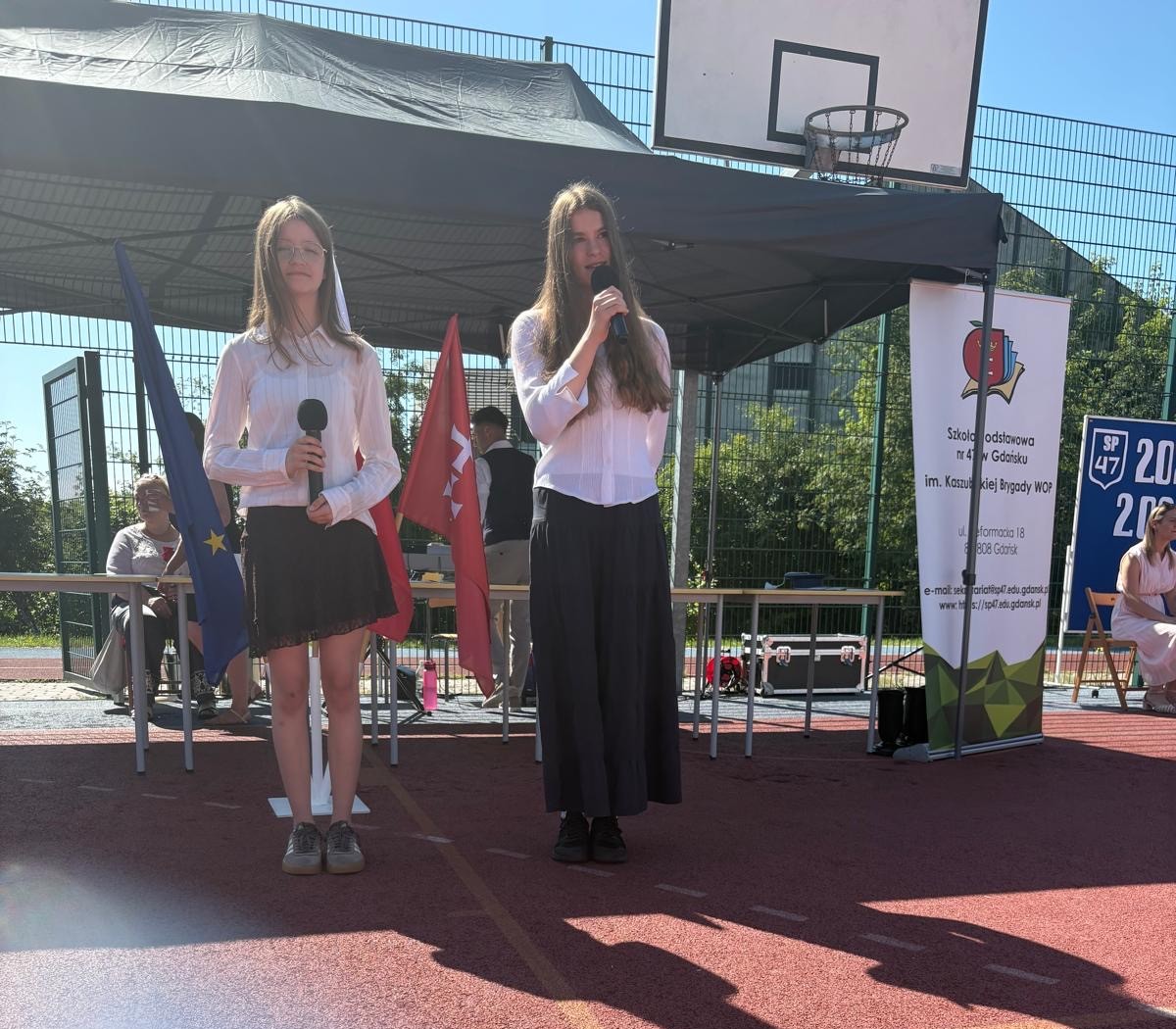  Dwie uczennice prowadzą część artystyczną uroczystości, stojąc na scenie z mikrofonami.