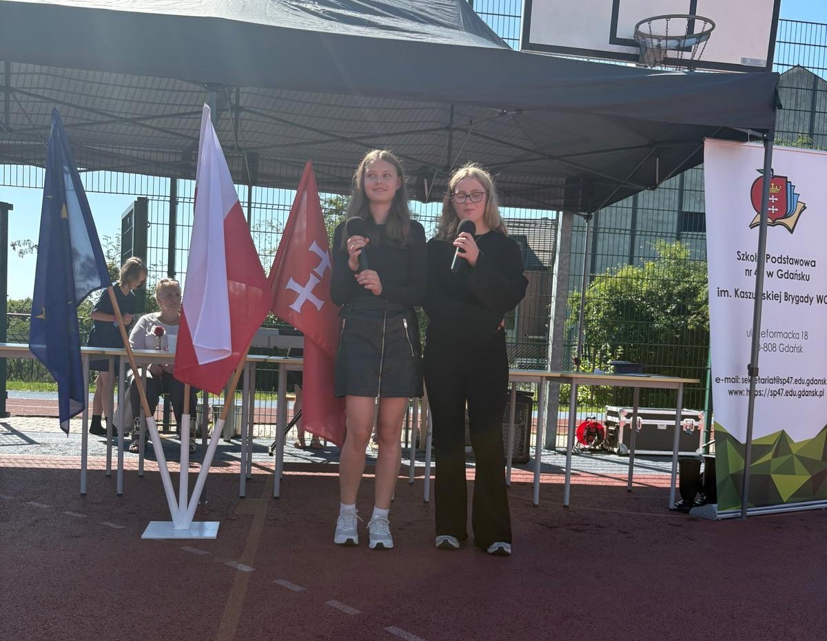Dwie uczennice występują na scenie z mikrofonami podczas szkolnej uroczystości zakończenia roku szkolnego.