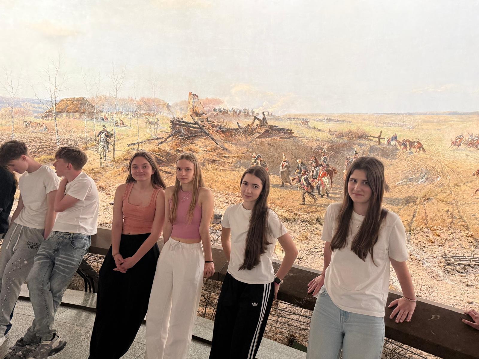 Cztery uczennice i dwóch uczniów znajduje się w muzeum historycznym. Dziewczęta stoją przy barierce i pozują do zdjęcia, natomiast dwóch chłopców po lewej stronie rozmawia ze sobą. W tle widoczna jest rozległa panorama przedstawiająca scenę bitwy z żołnierzami, zniszczonymi zabudo-waniami i dymem unoszącym się nad polem walki. Zdjęcie wykonano podczas zwiedzania ekspo-zycji muzealnej.