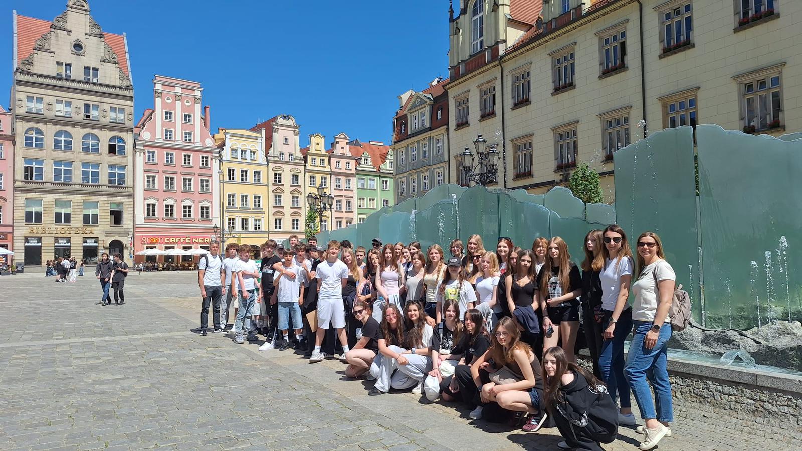 Grupa młodzieży i dwie dorosłe osoby pozują do wspólnego zdjęcia na rynku starego miasta we Wrocławiu w tle widać kolorowe kamienice, fontannę oraz bezchmurne, słoneczne niebo.