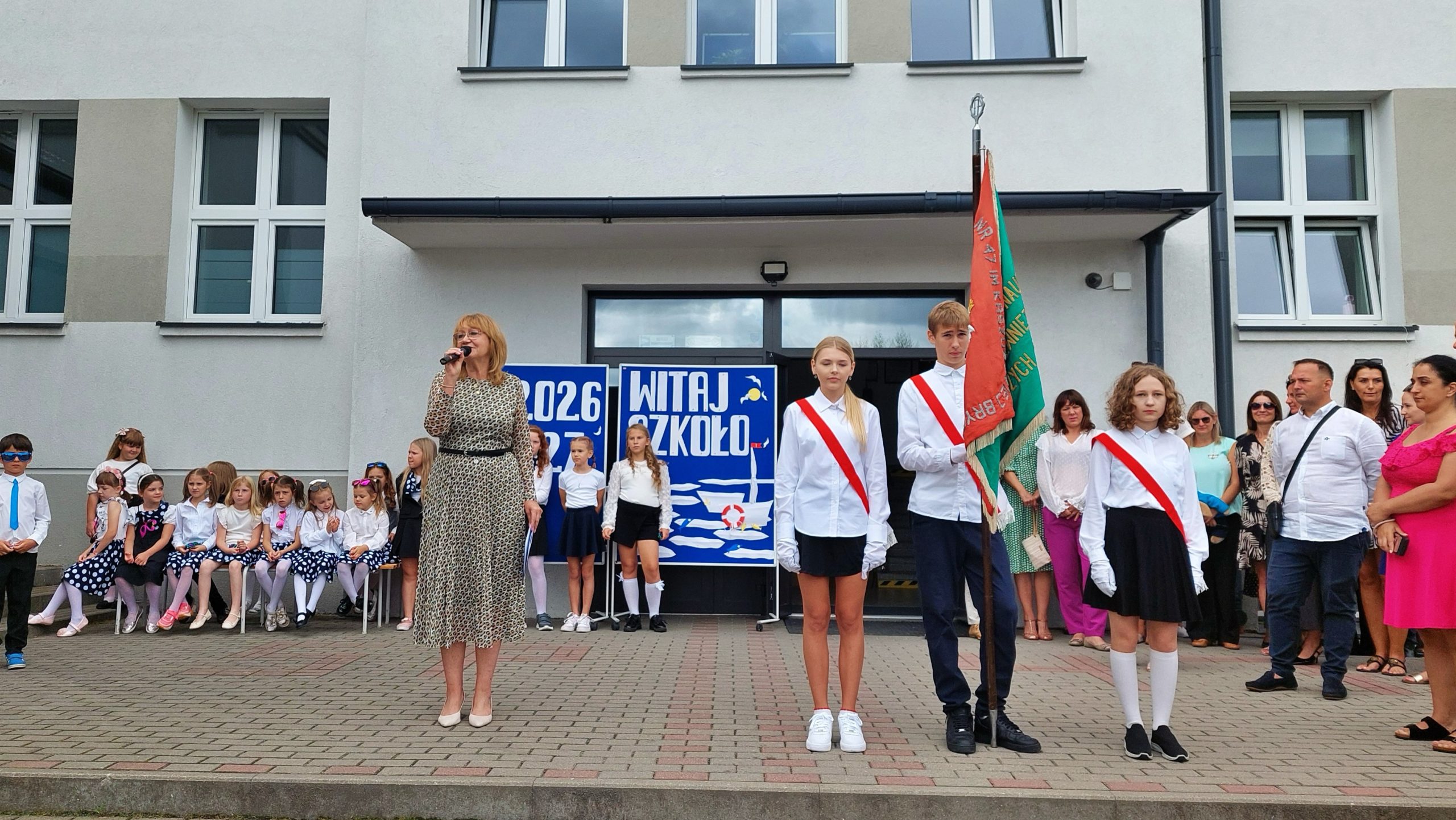 Przemówienie dyrektorki podczas rozpoczęcia roku szkolnego, obok niej uczniowie z pocztem sztandarowym i transparentem „Witaj Szkoło”